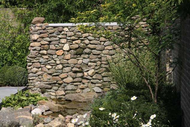 watermuur met opslagtank van 2500 liter water zodat er voldoende buffer aanwezig is. op de voorgrond een meerstammige solitaier struik gleditsia triacanthus hoogte muur 170 cm en breed 325 cm met maaskeien